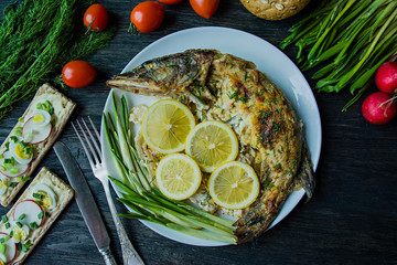 Baked pike in the oven, decorated with vegetables and herbs. Serving on a plate. Proper nutrition. Dark wood background.