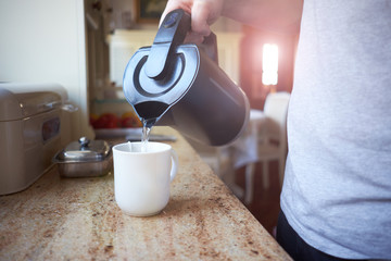 Hand of white man pouring hot boiling water from electric kettle to mug with tea bag