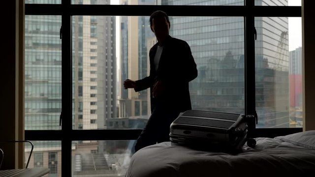 Man fiddle with small suitcase then throw it down on bed, waste time at hotel room. Guy idle around, silhouetted slow motion shot against bedroom window