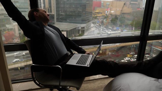 Man sitting with laptop, yawn and put hands on head, stretch body. Freelancer get tired of monotonous work, want to sleep, take short pause. City buildings and busy road seen outdoors