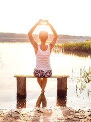 Fototapeta na wymiar A young girl sits on a bench by the river enjoys a beautiful sunset and shows hands fingers heart sign. Girl sitting near water outdoors. Golden sunset on the lake. Young woman thinking about