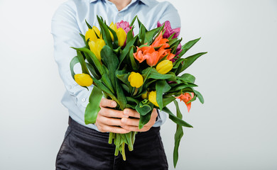 The man is holding in hands colorful tulips on a light background. The concept of handing flowers to a woman, girl. A man wearing a shirt is holding flowers in his hands.