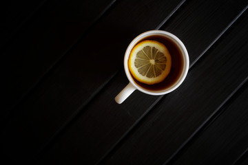 A white cup with black tea and a slice of lemon stands on a dark wooden table. View from above. Minimalism