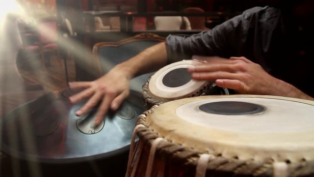 A man playing an ethnic musical instruments tabla and hang in the interior of the lounge, with the rays of the sun from window