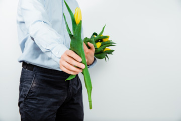 The man is holding in hands yellow tulips on a light background. The concept of handing flowers to a woman, girl. A man wearing a shirt is holding flowers in his hands.