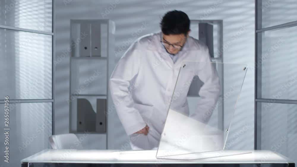 Poster middle eastern male doctor in lab coat entering office, turning on desk lightning and typing on futu