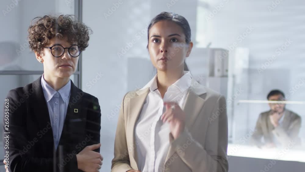 Poster caucasian and asian businesswomen tapping and swiping on invisible multi-touch screen on glass wall 