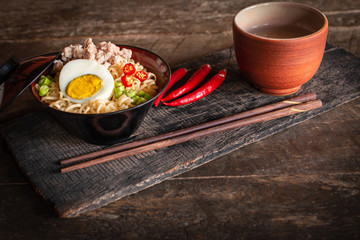 Fototapeta na wymiar Instant noodle with pork, egg and vegetables on black bowl on the wood table there are chilli, chopstick and drinking glass placed around.