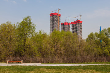 Three high-rise buildings under construction. In the foreground is a park and a street bench.