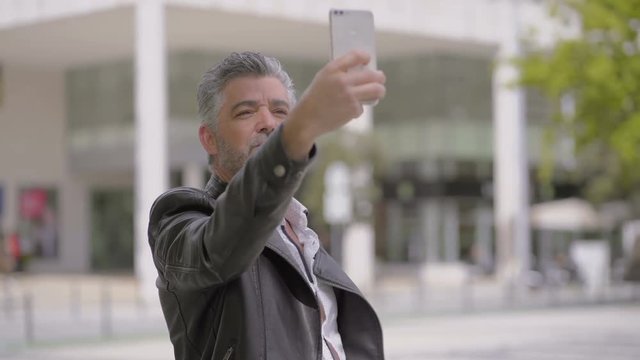 Mature man taking selfie with smartphone on street. Handheld shot of stylish middle aged man in leather jacket taking pictures with cell phone and smiling outdoor. Technology concept