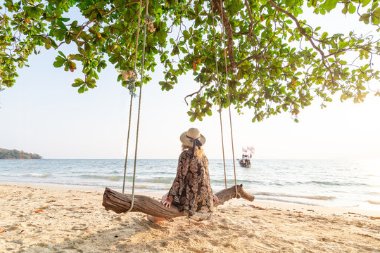 Young blonde woman sitting on a wooden swing at Krabi Railey beach overlooking the harbour and long tail boat, Thailand