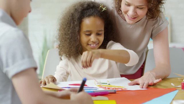 Cute little African girl smiling and using scotch tape while making creative painting with help of female teacher in art class
