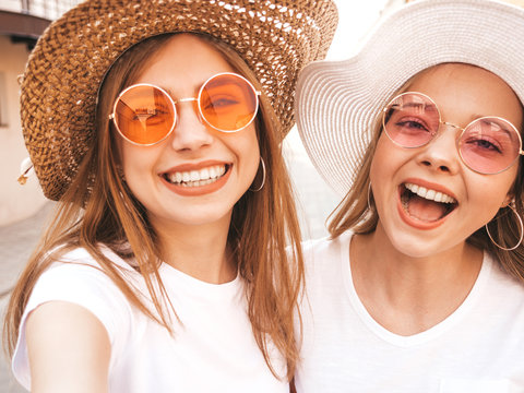 Two young smiling hipster blond women in summer white t-shirt clothes. Girls taking selfie self portrait photos on smartphone.Models posing on street background.Female showing positive face emotions