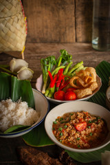 Thai Northern Style Pork and Tomato Chili Relish, nam prik ong in white bowl on wood table there are side dishes of fresh vegetables, cooked rice, water bowl, rice wicker and flower placed around.