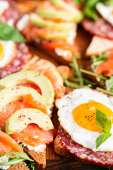 Huge healthy breakfast with sun-dried tomatoes, sandwiches with scrambled eggs, sausage, salmon, arugula, curd cheese, avocado on a wooden board