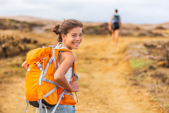 Happy young Asian hiker girl hiking with friend in mountain nature trail wearing orange backpack smiling looking back enjoying travel holiday in summer. Adventure wanderlust lifestyle. Healthy woman.