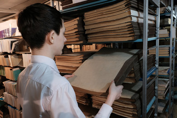 A schoolboy in a white shirt pulls out an old book from a bookshelf. A student in the library or in the archive room is looking for information. Concept back to school or study of historical documents