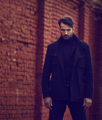 Frustrated evil young man in black coat near the brick wall looking strong. Closeup toned portrait