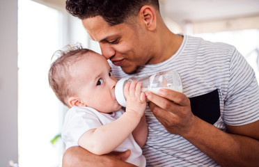 A portrait of father bottle feeding a small toddler son indoors at home.