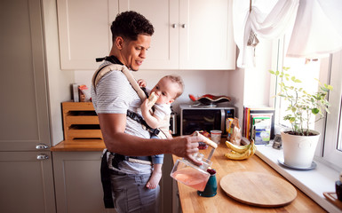 A father with small toddler son in carrier in kitchen indoors at home.