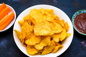 Crispy potato chips in a bowl with carrot slices and tomato sauce on a textured background.