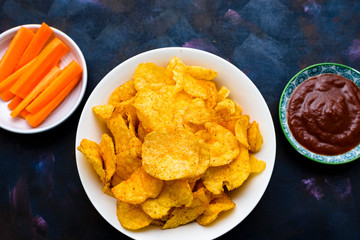 Crispy potato chips in a bowl with carrot slices and tomato sauce on a textured background.