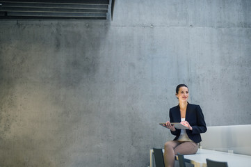 Young business woman with tablet sitting on desk against concrete wall in office.