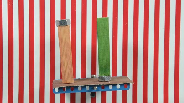 Two alike wood blocks with a metal at one end paced on a board that tgradually tilts, one by one they fall. The background lines are vertical reference, 