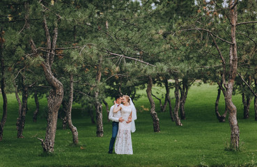 Happy newlyweds tenderly embracing in the forest against the background of green grass and pines. Wedding portrait of a stylish groom in a shirt and a cute curly bride in a white dress.
