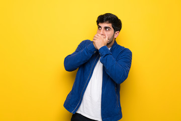 Man with blue jacket over yellow wall covering mouth and looking to the side