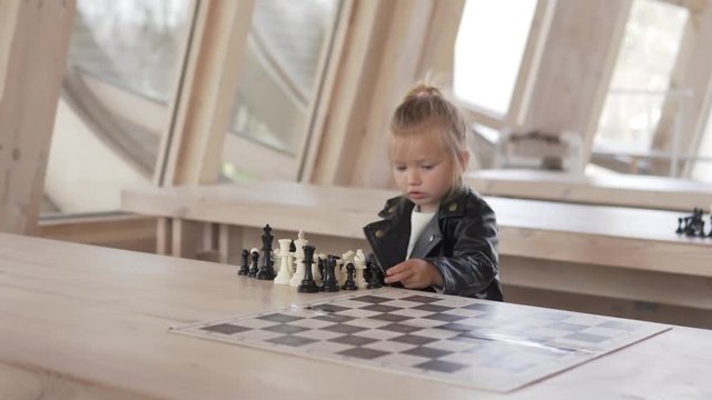 Beautiful little girl plays with chess pieces. She sits at a wooden table in a chess club and removes pieces from a game board.
