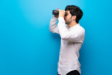 Young man over isolated blue wall and looking in the distance with binoculars