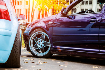 Two modified low cars in purple candy and light blue color. Stance custom cars with a forged polished wheels parked on a street at sunny day. Tuned automobiles