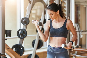 Fototapeta premium Exercising with dumbbells. Sporty young woman in sportswear and with smartwatch on her hand exercising with dumbbells while standing at gym