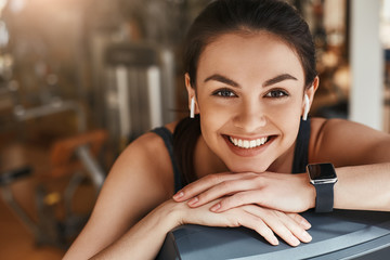 Always in good mood! Portrait of cute and cheerful woman with smart watch on her hand looking at camera with toothy smile while taking a rest at gym.