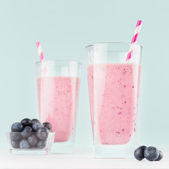 Two healthy dairy desserts of ripe blueberry in glass with berries in bowl, striped straws on white wood board and pastel green mint background, square.