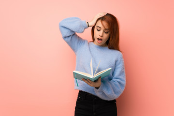 Young redhead woman over pink background surprised while enjoying reading a book