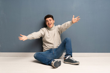 Young man sitting on the floor presenting and inviting to come with hand
