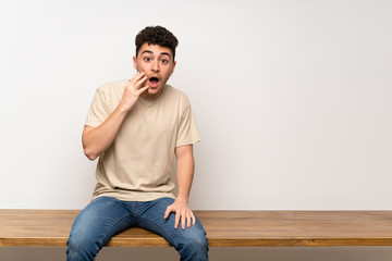 Young man sitting on table with surprise and shocked facial expression