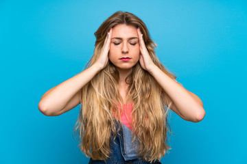 Young blonde woman with overalls over isolated blue wall with headache