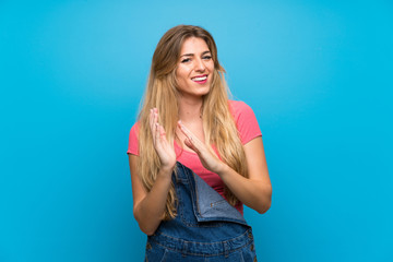 Young blonde woman with overalls over isolated blue wall applauding after presentation in a...