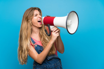 Young blonde woman with overalls over isolated blue wall shouting through a megaphone