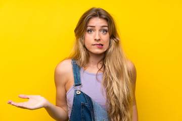 Young blonde woman with overalls over isolated yellow background having doubts and with confuse face expression