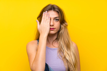 Young blonde woman with overalls over isolated yellow background covering a eye by hand