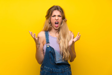 Young blonde woman with overalls over isolated yellow background unhappy and frustrated with something