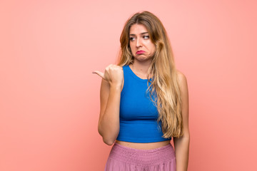 Young blonde woman over isolated pink background unhappy and pointing to the side