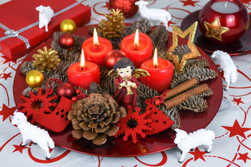 Red Christmas decorated wreath plate with four burning candles on a table cloth surrounded with white sheep and other Xmas theme decoration