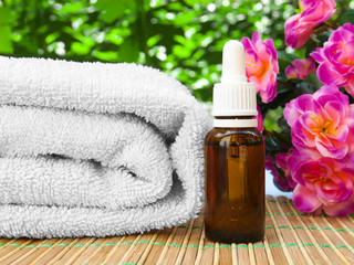 Obraz na płótnie Canvas Floral spa and wellness concept. Aromatic oil bottle on a bamboo placemat with white towel. Purple flower and blurred green plants in the background. Outdoors with shallow depth of field, bokeh effec.