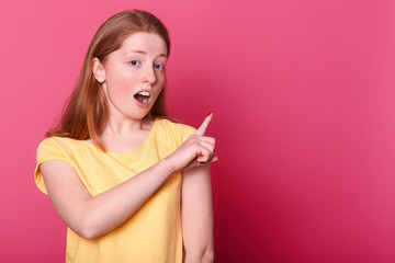Indoor shot of charismatic attractive sweet young girl, shows direction aside, looks impressed, opening mouth and eyes widely. Sincere red haired model poses over bright pink background in studio.
