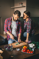 Beautiful young couple in kitchen at home while cooking healthy food. Man is cuts salad. Woman sits on table and he looks at her husband. Scene from family life.